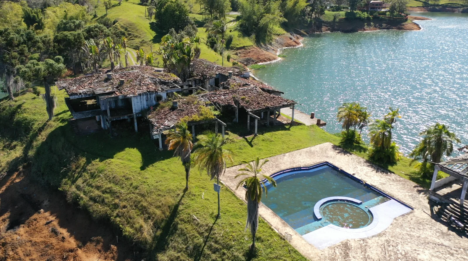 Close aerial view of the ruined main house at Finca La Manuela with collapsed tile roofs, roofless walls and a columned portico, beside the estate's empty swimming pool and the reservoir